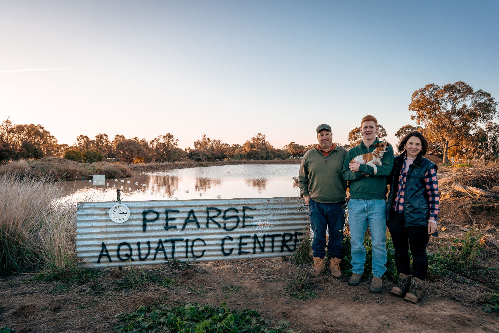 Paralympian Col Pearce and his parents standing in front of family farm dam with a sign which reads "Pearce Acquatic Centre"
