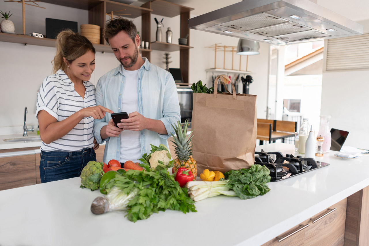 Image of man and woman standing in kitchen with groceries on the bench, looking at mobile phone screen. 