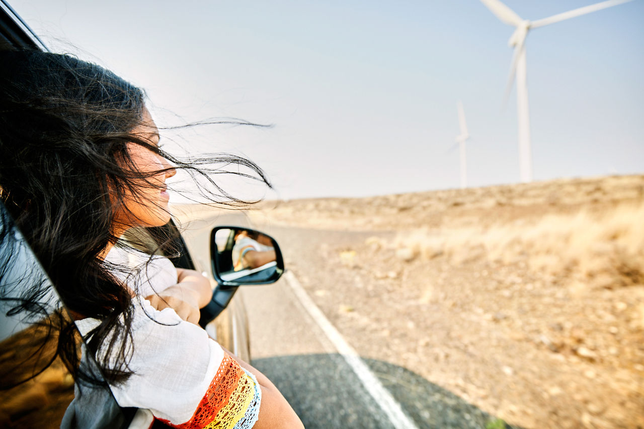 Smiling teenage girl with head out car window and hair blowing in wind passing wind turbines in dessert